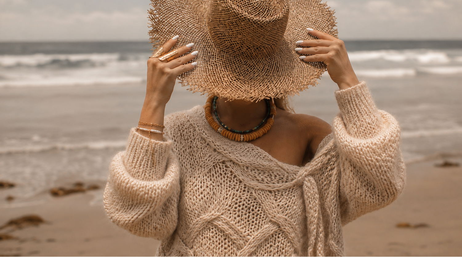 A woman on a beach wearing a large woven straw sun hat, a chunky knit cream sweater, and a beaded necklace, holding the brim of the hat with both hands.